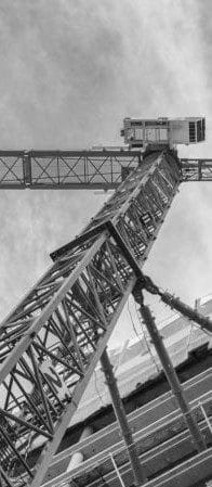 Ein Schwarzweißfoto eines Baukrans, der über einem im Bau befindlichen Gebäude aufragt, von unten vor einem bewölkten Himmel betrachtet, unterstreicht die Bedeutung von Vibrationsschutz und Schwingungsisolation in modernen Bauwerken.