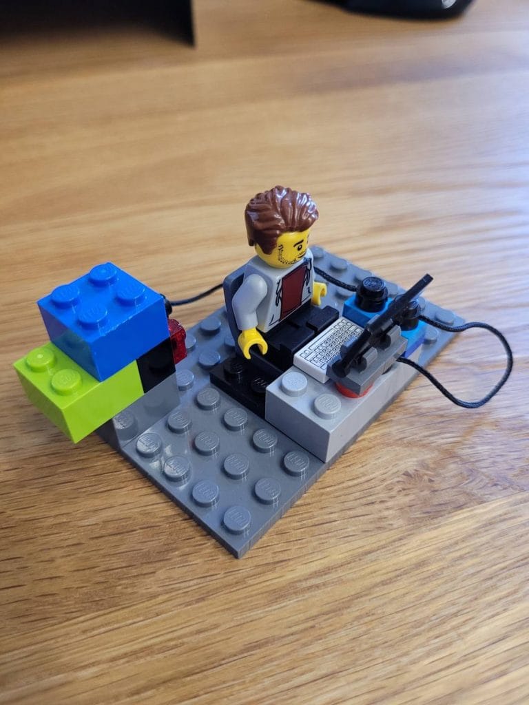 Ein LEGO Bauingenieur sitzt an einem Schreibtisch mit einem Computer, einer Tastatur und einem Headset, verbunden mit einer blockartigen blauen und grünen Struktur auf einer Holzoberfläche.