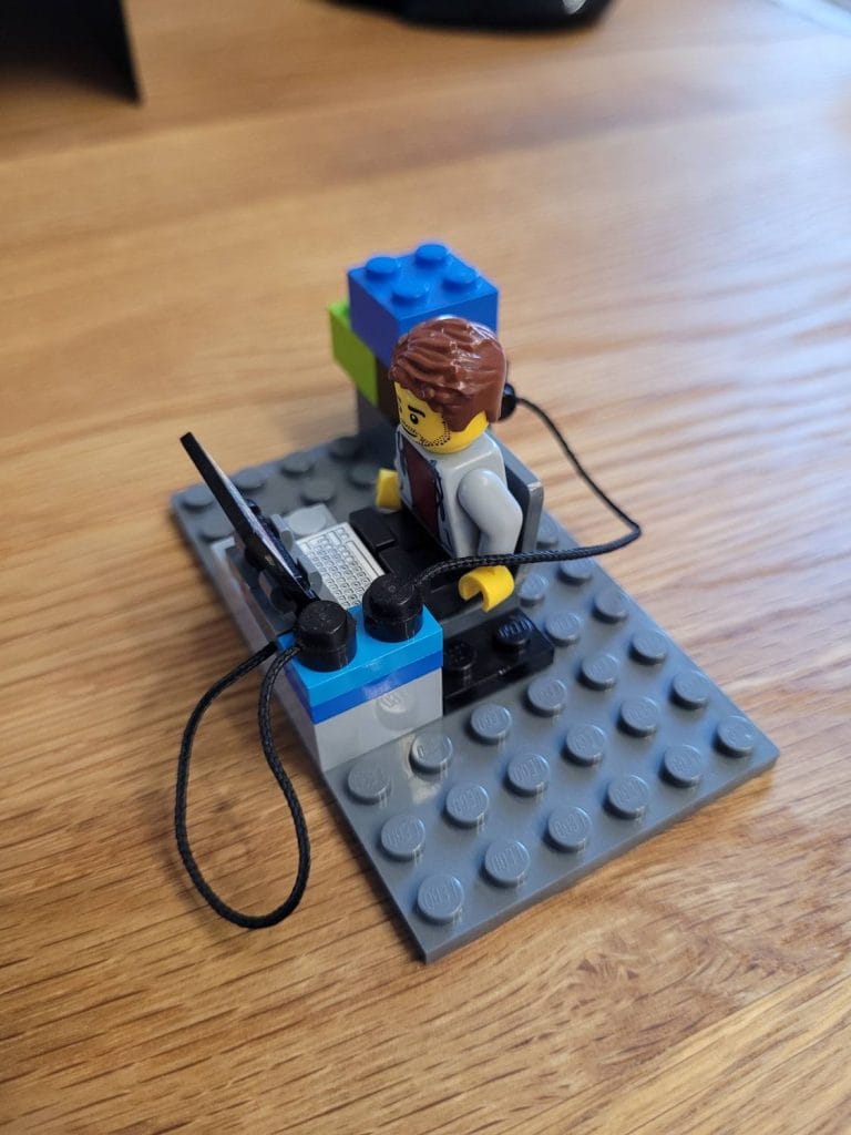 Ein LEGO Bauingenieur, der an einem grauen Schreibtisch mit einem Computer, zwei Antennen und Kabeln auf einer Holzoberfläche sitzt.