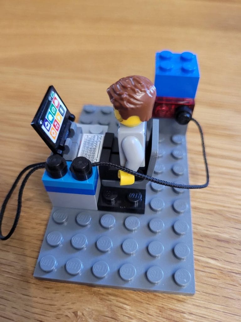 Ein LEGO Bauingenieur, der an einem Schreibtisch mit einem Laptop sitzt, umgeben von blauen Blöcken und schwarzen Kabeln auf einer grauen LEGO Grundplatte.