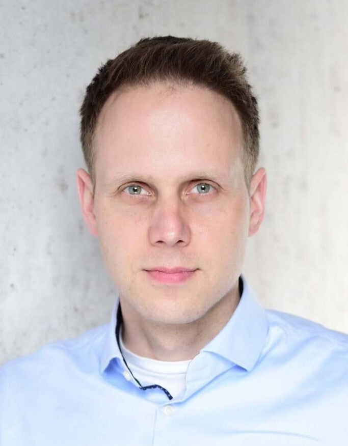 Ein Mann mit kurzen braunen Haaren und hellen Augen trägt ein hellblaues Hemd mit Kragen und schaut vor einem neutralen Hintergrund direkt in die Kamera.