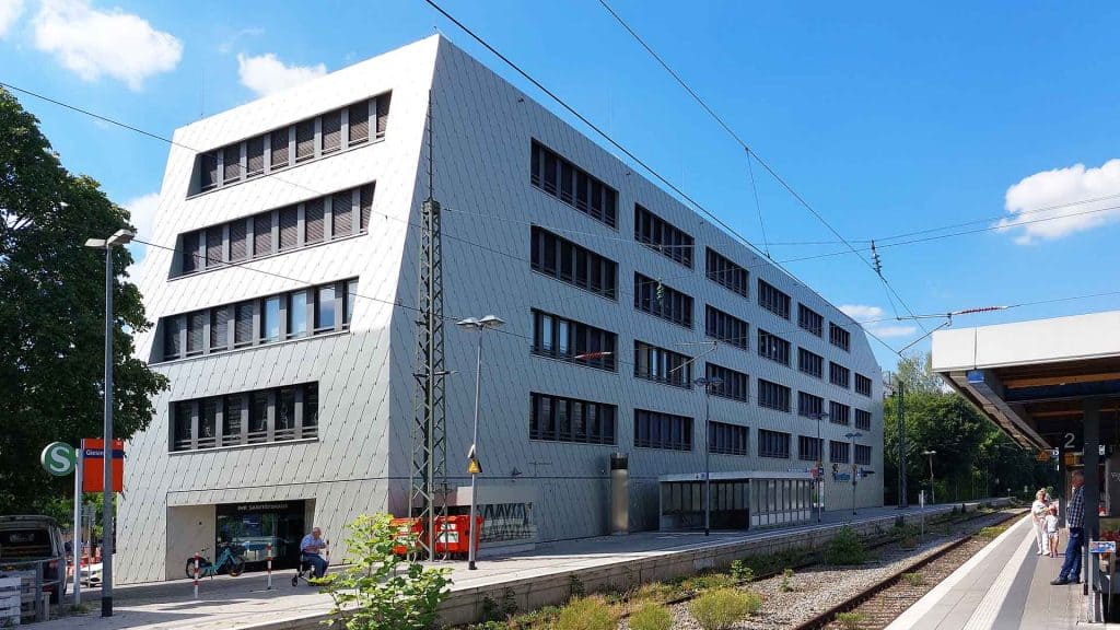Modernes, kantiges, silbernes Gebäude mit unregelmäßiger Fensteranordnung neben einem Bahnsteig unter blauem Himmel, mit elastischer Gebäudelagerung für erhöhten Schwingungsschutz. Elastische Gebäudelagerung