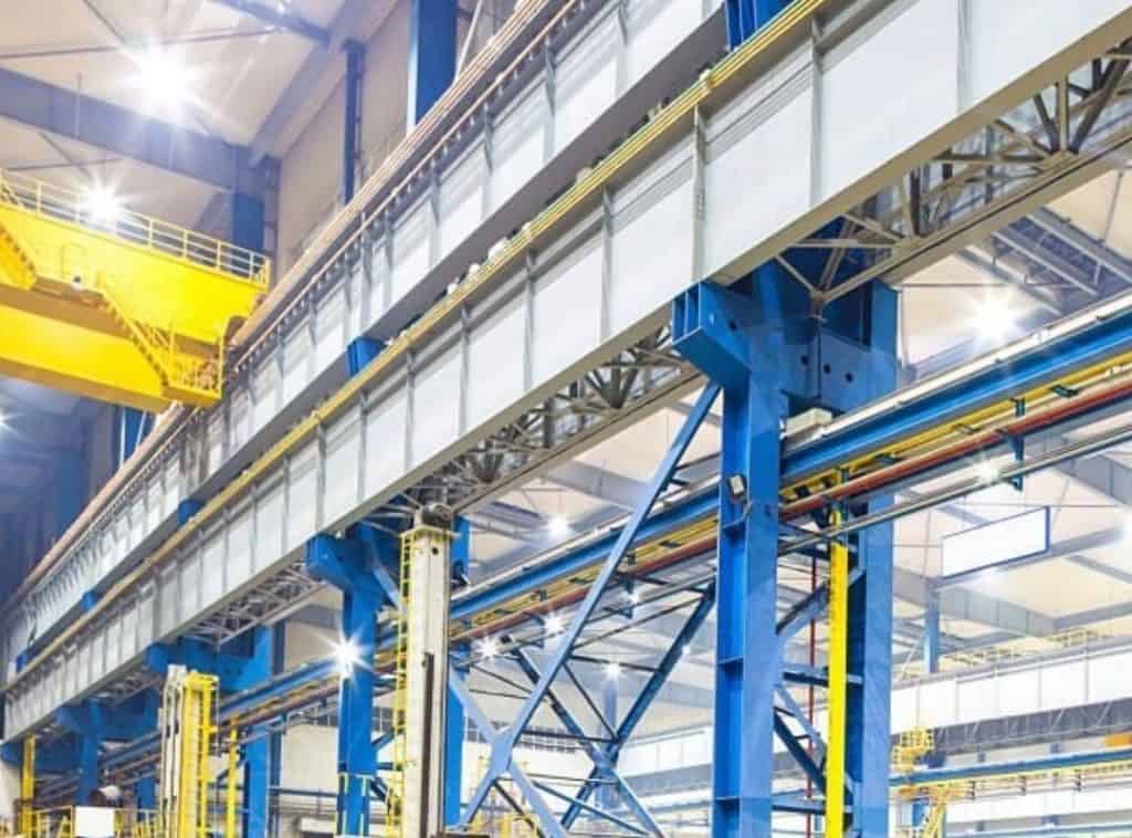 Innenraum einer großen Industrieanlage mit blauen Stahlträgern, gelben Geräten, Brückenkränen und Metallstegen unter heller Beleuchtung, konzipiert für elastische Entkopplung zur Reduzierung von Erschütterungen im gesamten Arbeitsbereich. Stahl- und Industriebauten