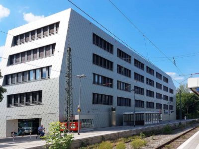 Ein modernes fünfstöckiges Gebäude mit geometrischen Fenstern und innovativer Anwendungstechnik steht neben einem Bahnsteig unter strahlend blauem Himmel; ein paar Menschen warten auf dem Bahnsteig.