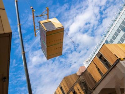 Ein Kran hebt ein großes vorgefertigtes Gebäudemodul über eine Baustelle, auf der moderne Gebäude und innovative Anwendungstechnik unter einem teilweise bewölkten Himmel präsentiert werden.