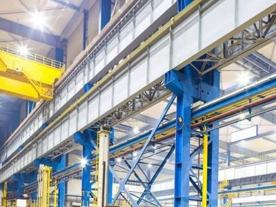 Das Innere einer großen Industrieanlage mit blauen Stahlträgern, Brückenkränen und verschiedenen Rohren und Kabeln, die entlang der Struktur verlaufen, ist für effiziente Anwendungstechnik ausgelegt.