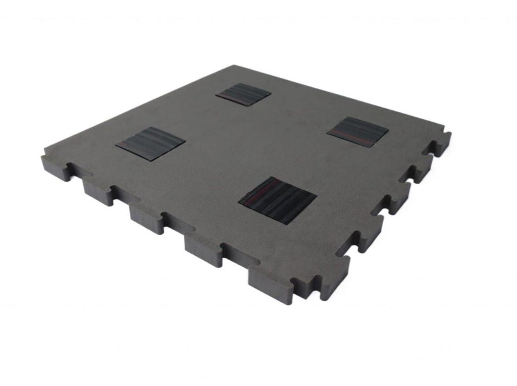 Dunkelgraue ineinandergreifende Bodenfliese mit vier schwarzen Quadraten auf der Oberfläche, fotografiert vor einem weißen Hintergrund.